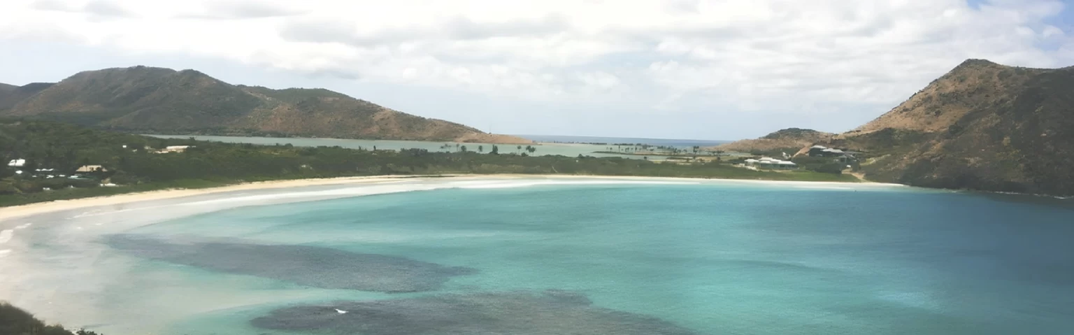 Turquoise bay with sandy shoreline, surrounded by green hills under a partly cloudy sky.