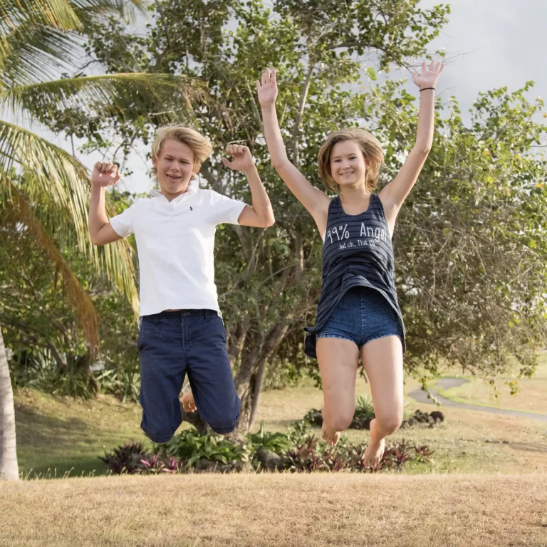 Two kids joyfully jumping with arms raised in a sunny park setting with lush greenery.