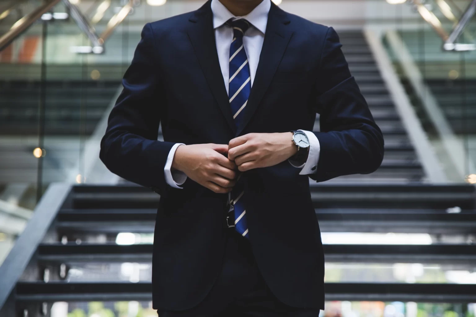 Man in a suit adjusting his jacket, standing confidently in front of a staircase.