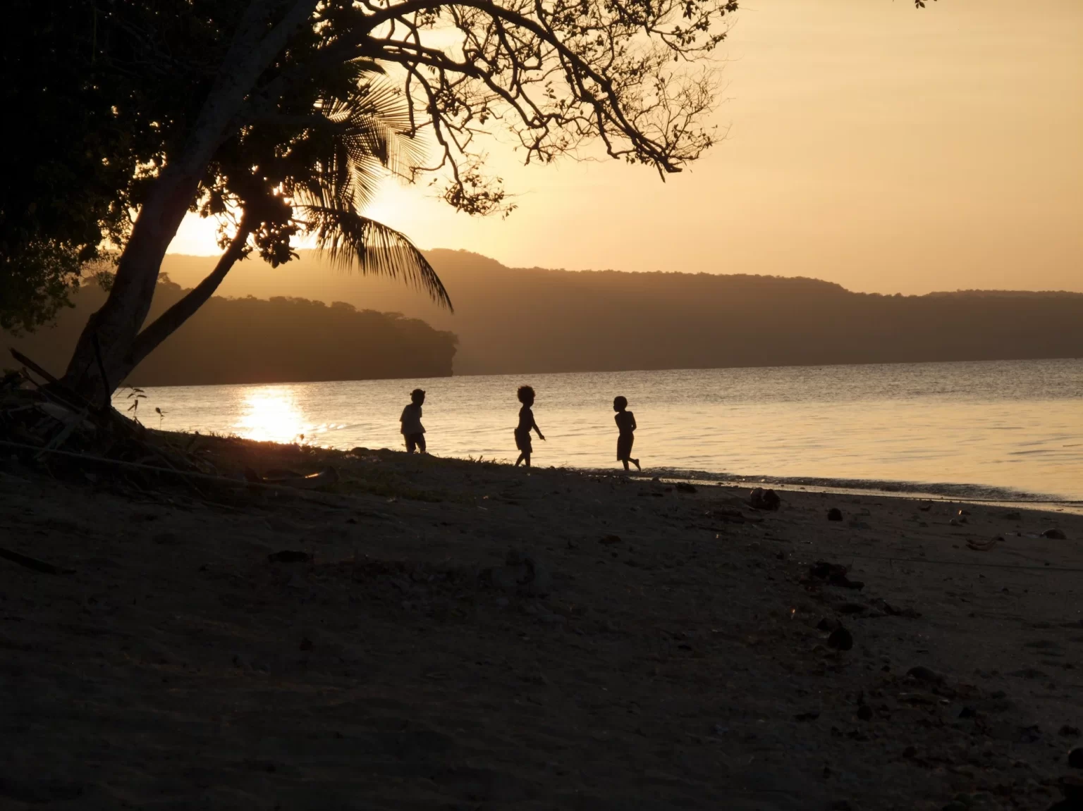Three children walking along a beach at sunset, silhouetted against a bright horizon.