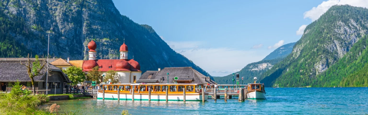 Boat docked near St. Bartholomew's Church with red domes, set against a backdrop of alpine mountains and a clear blue lake.