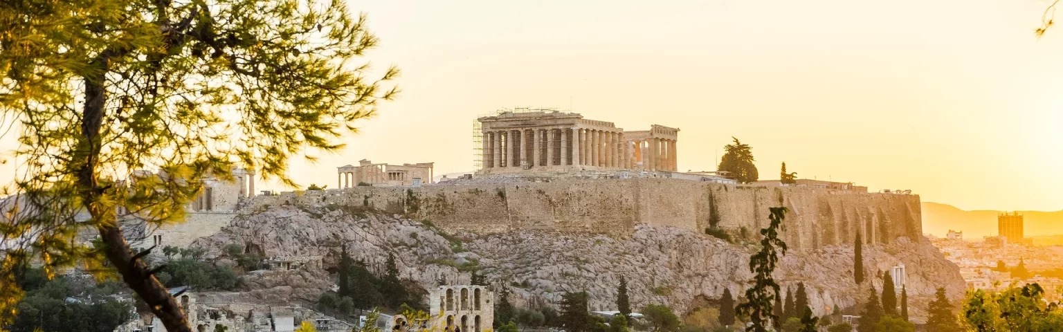 The Acropolis in Athens at sunset with the Parthenon visible atop a rocky hill, surrounded by trees.