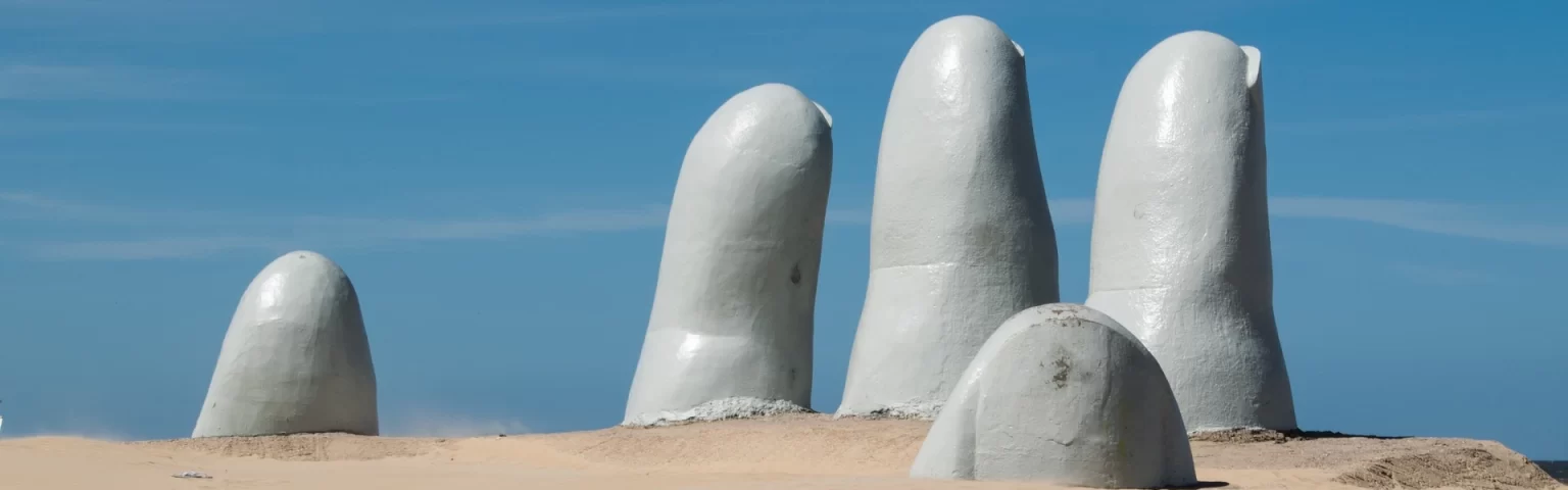Giant concrete fingers protrude from sandy beach under a clear blue sky.