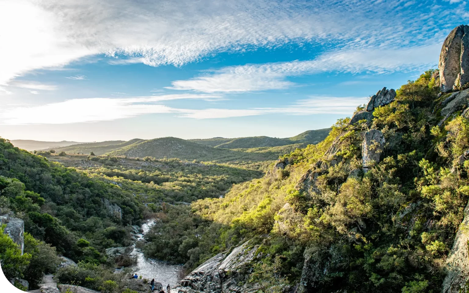 Scenic view of a lush valley with rolling hills, a flowing stream, and a bright blue sky.