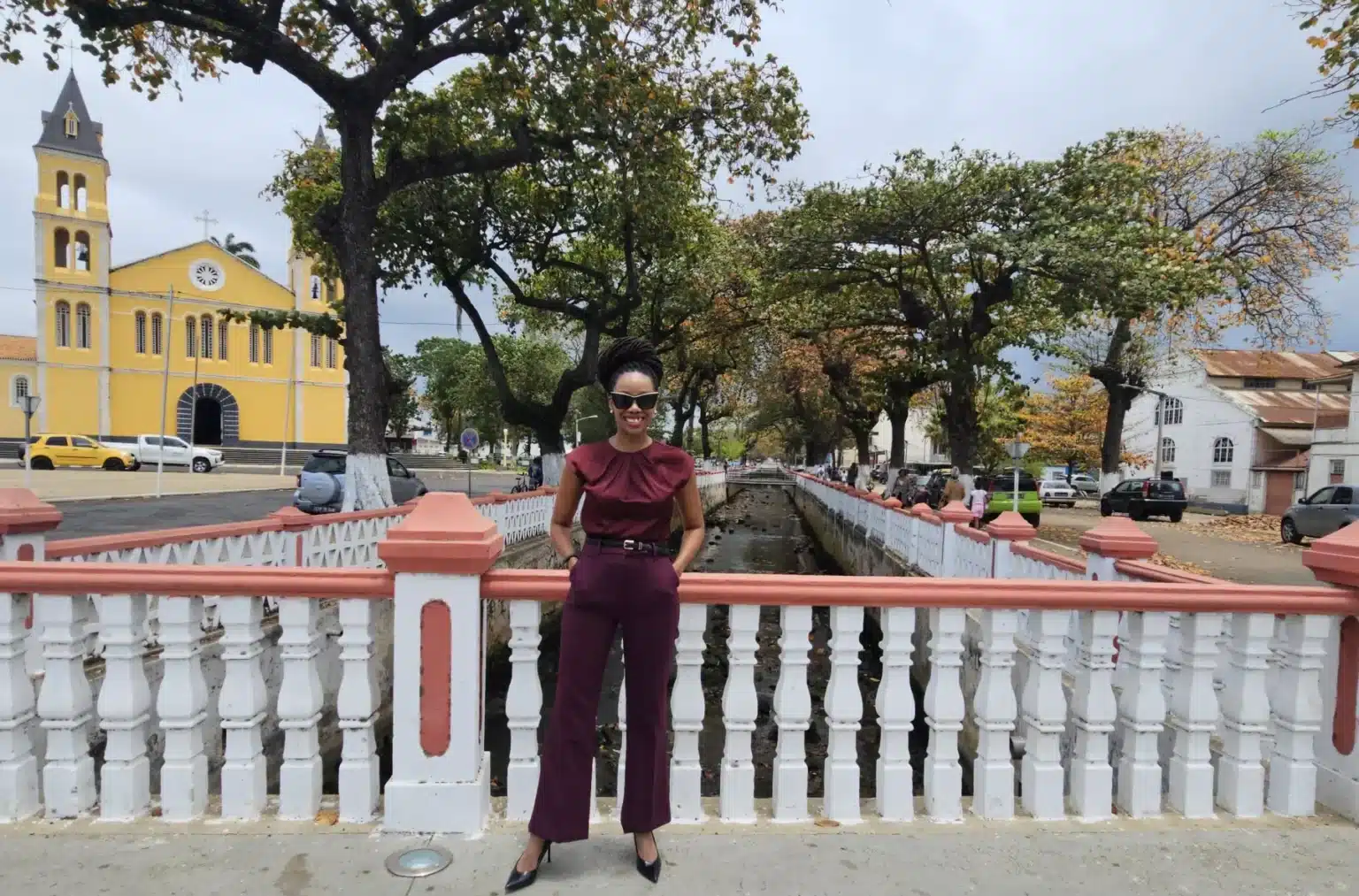 Angela Blair Cadet standing on a pedestrian bridge with a yellow church in the background in São Tomé and Príncipe.